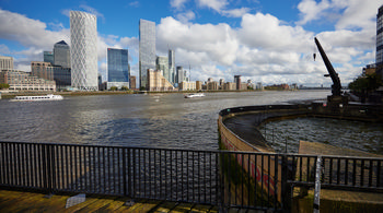 Canary Wharf Dock Crane 2 This urban landscape photograph shows Canary Wharf Dock Crane 2 located on the banks of the River Thames in London, United Kingdom. The image was taken in the afternoon during early autumn, as indicated by the partly cloudy sky and bright sunlight, which highlight the modern skyline of Canary Wharf with its distinctive high-rise buildings, including the HSBC Tower and Newfoundland Tower. Several boats are visible on the River Thames, adding to the bustling city atmosphere. The photograph captures the site's unique mix of historical dockside elements and contemporary architecture found in this prominent financial district of London.